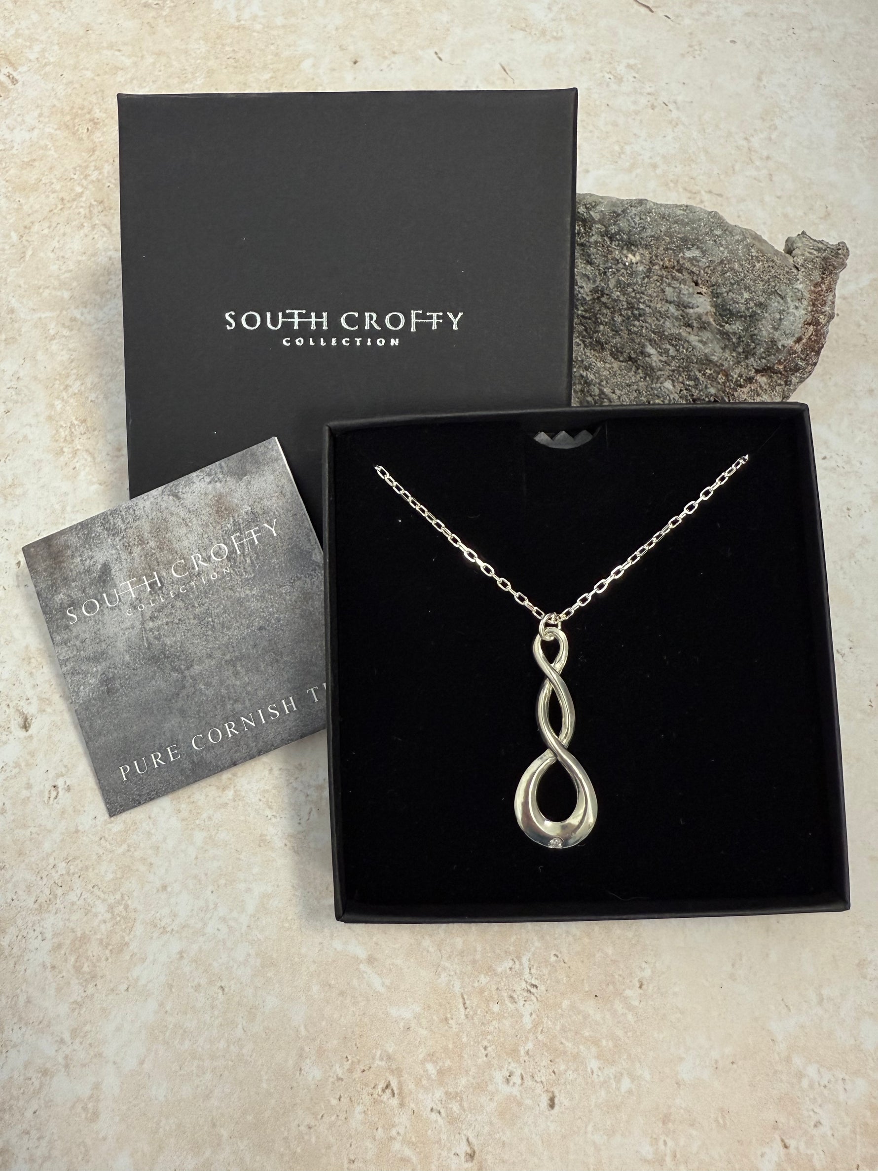 A Cornish tin pendant with a double twist design and a central sparkling diamond, presented on a 18-inch chain. In a black South Crofty presentation box with a certificate of authentication on a grey background.