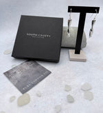Cornish tin earrings with diamonds on a stand next to a South Crofty branded box and card on a textured surface.