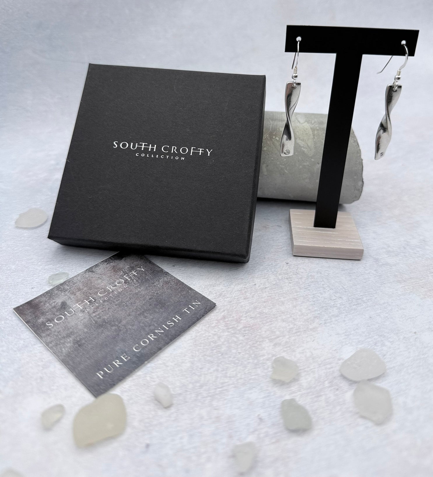 Cornish tin earrings with diamonds on a stand next to a South Crofty branded box and card on a textured surface.