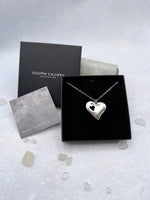 Silver heart-shaped necklace with cutout heart and a diamond in a South Crofty box on a light gray background