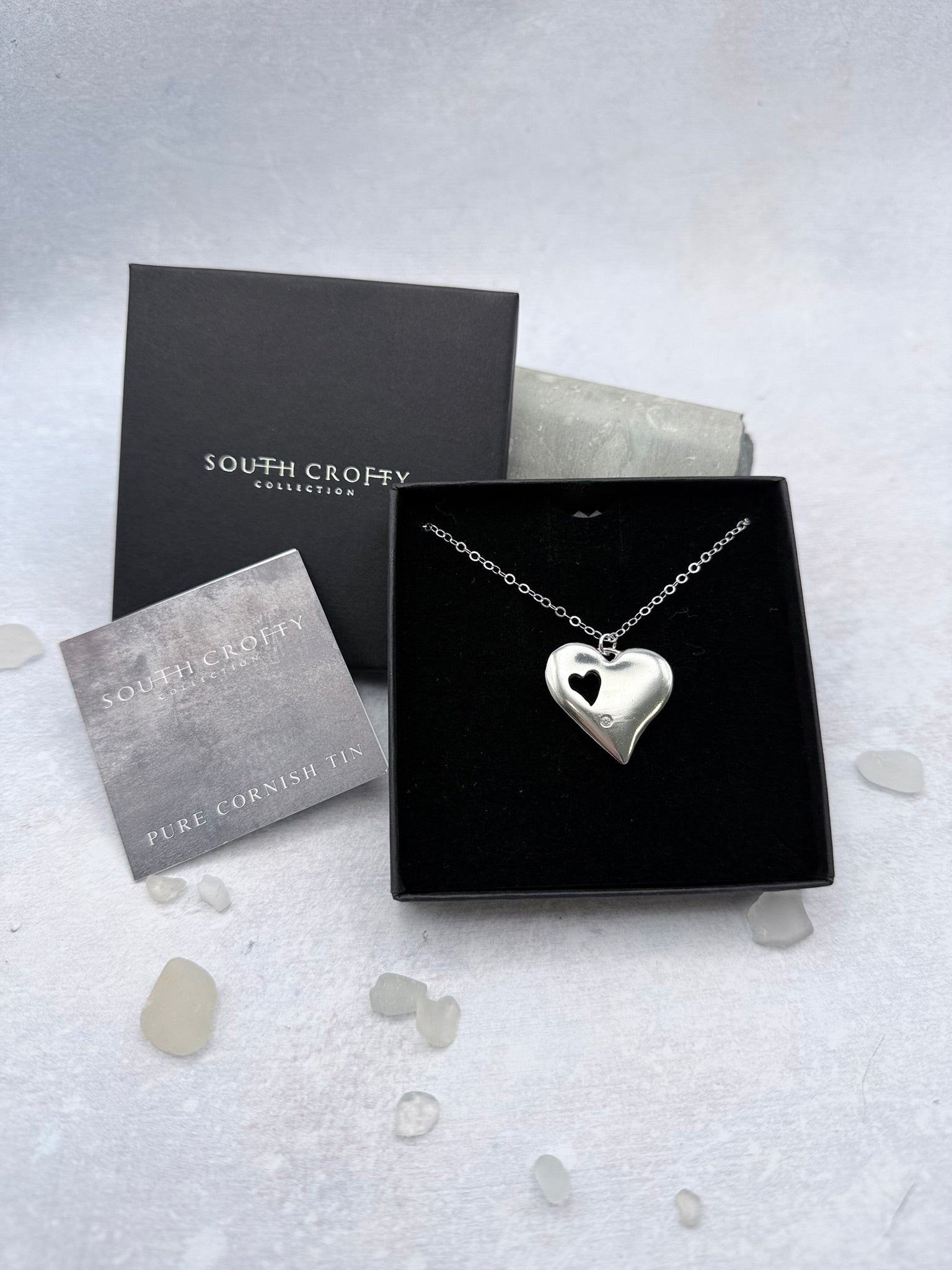 Silver heart-shaped necklace with cutout heart and a diamond in a South Crofty box on a light gray background