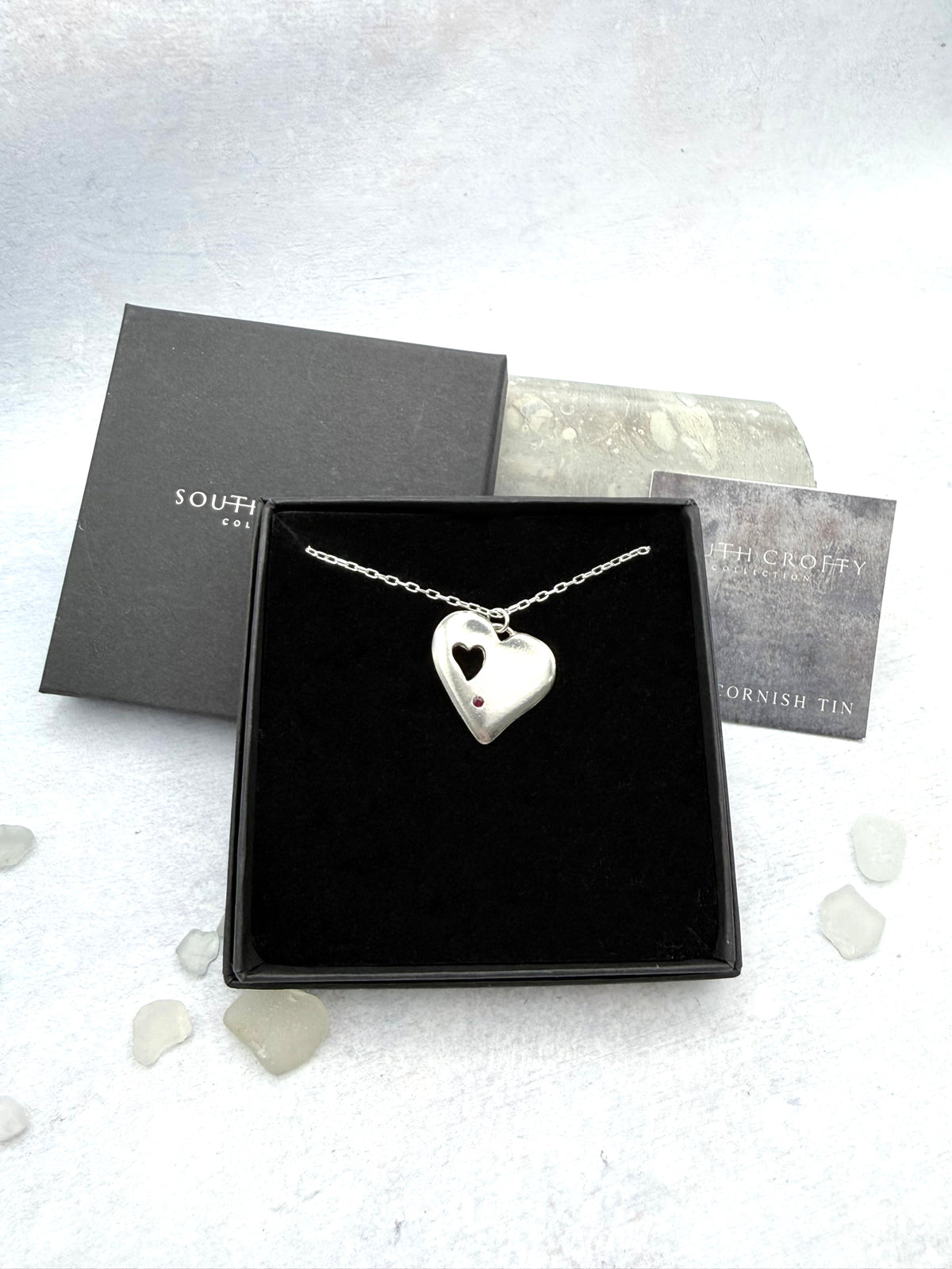 Cornish tin heart-shaped pendant with cut-out and a ruby gemstone in a black box with 'South Crofty' branding on a light grey background.