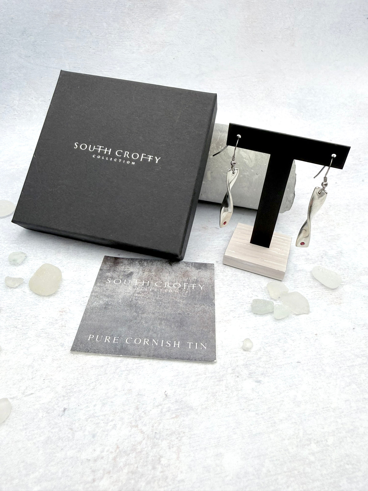 Cornish tin earrings with a twist design and inset rubies hanging beside a South Crofty branded presentation box and certificate of authenticity