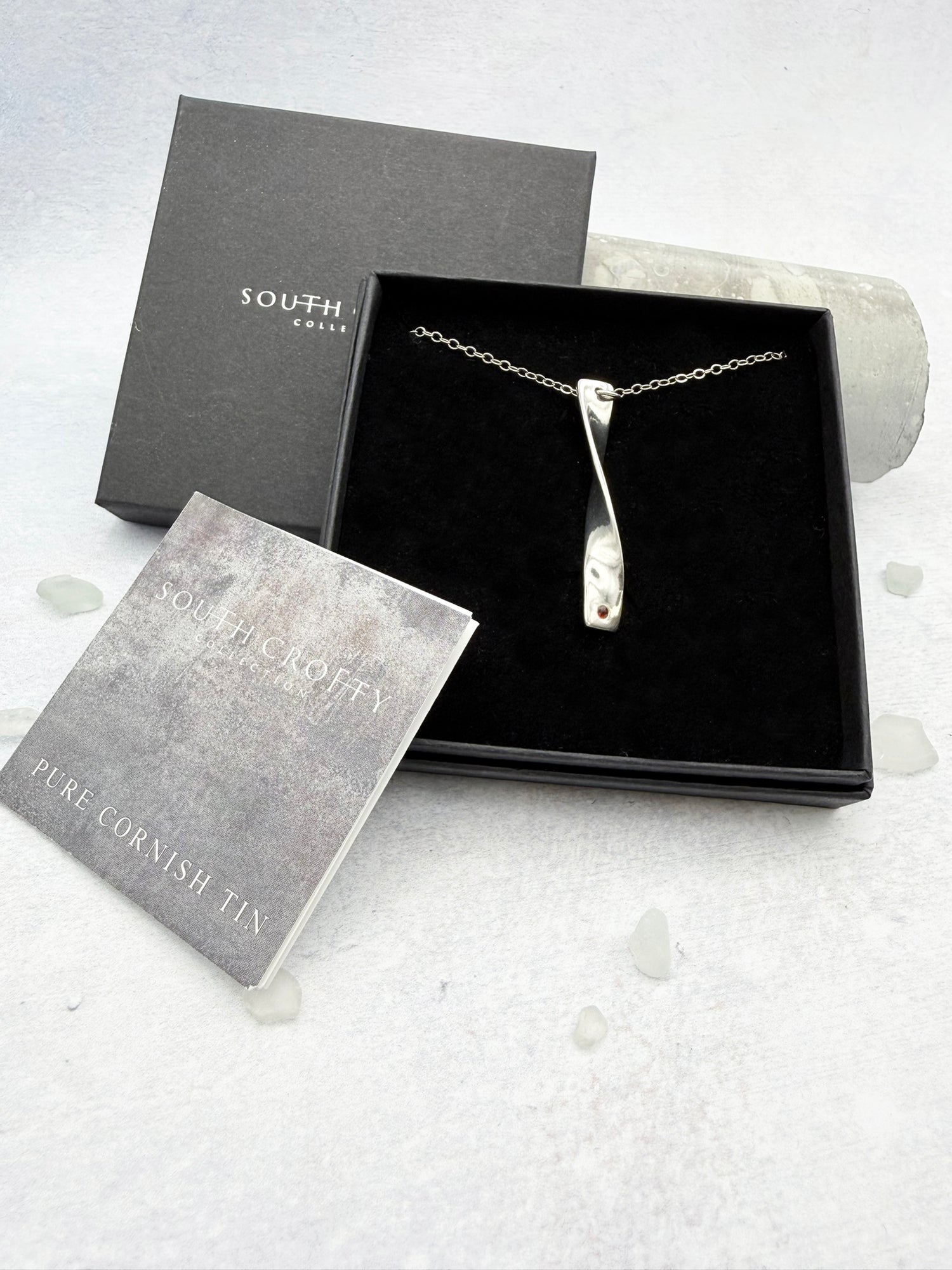 Cornish tin necklace which is silver in colour in a box with a certificate of authenticity for the tin on a light gray background