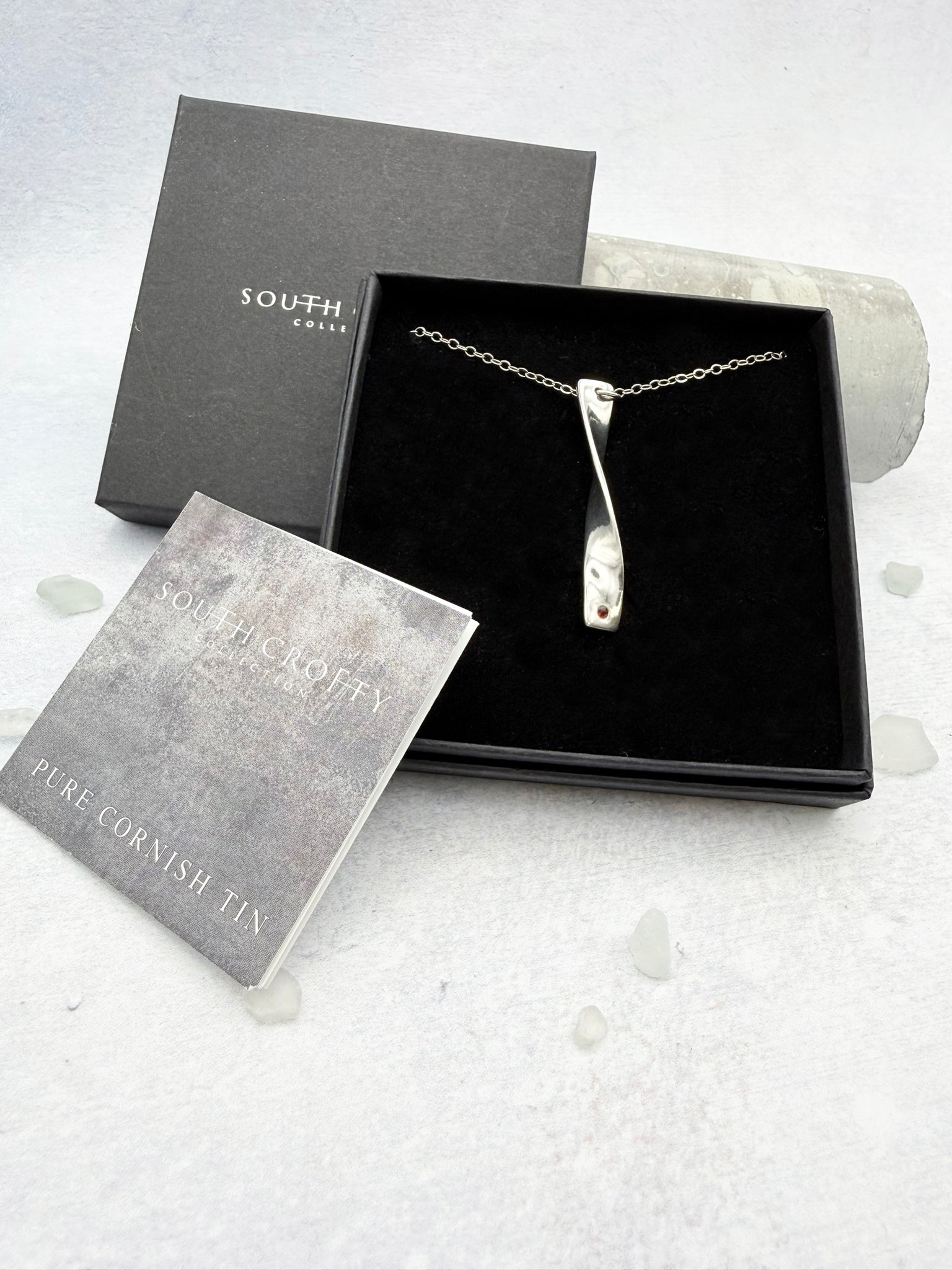 Cornish tin necklace which is silver in colour in a box with a certificate of authenticity for the tin on a light gray background