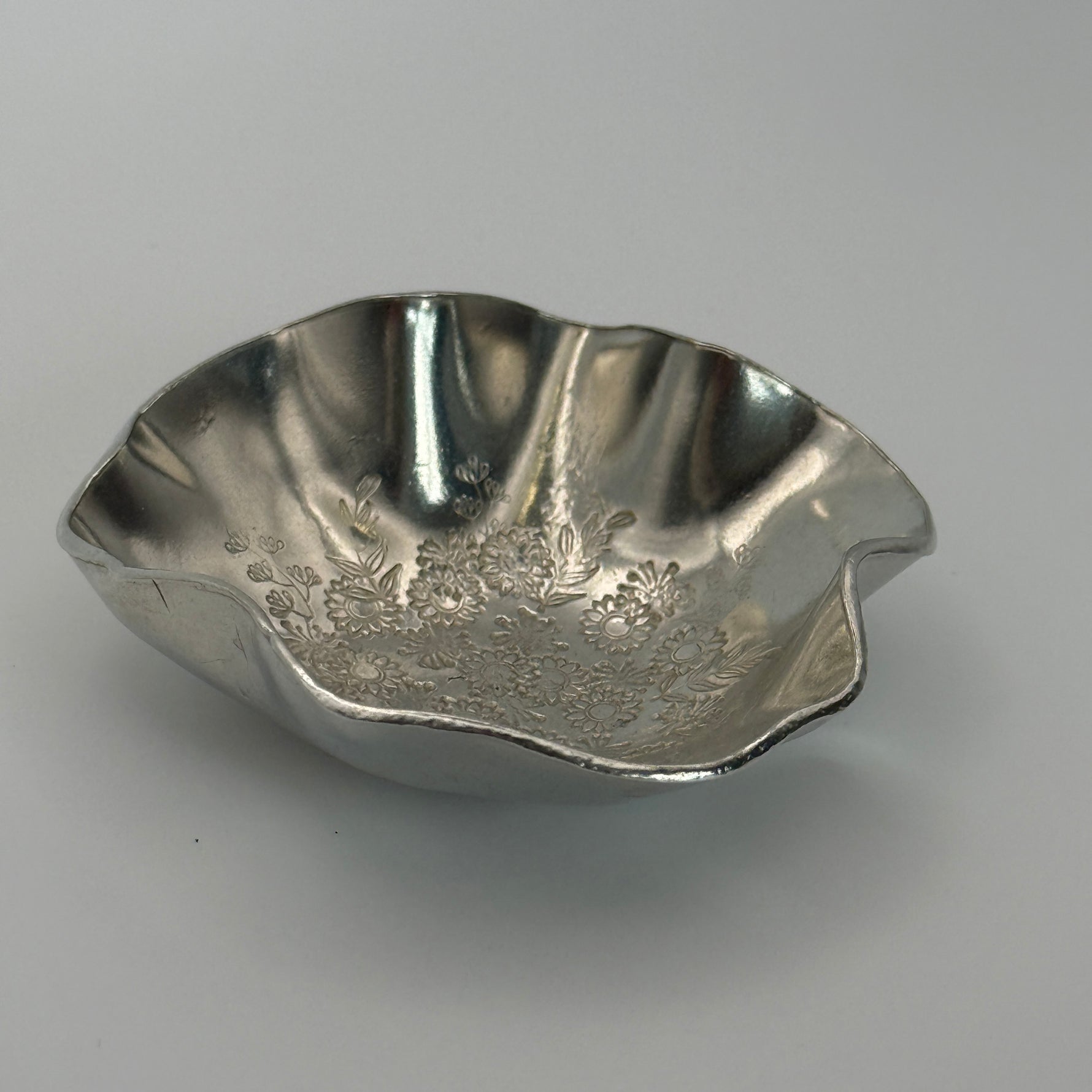 A trinket dish handmade from Cornish tin with waved edges and a hand stamped flower design in the base.